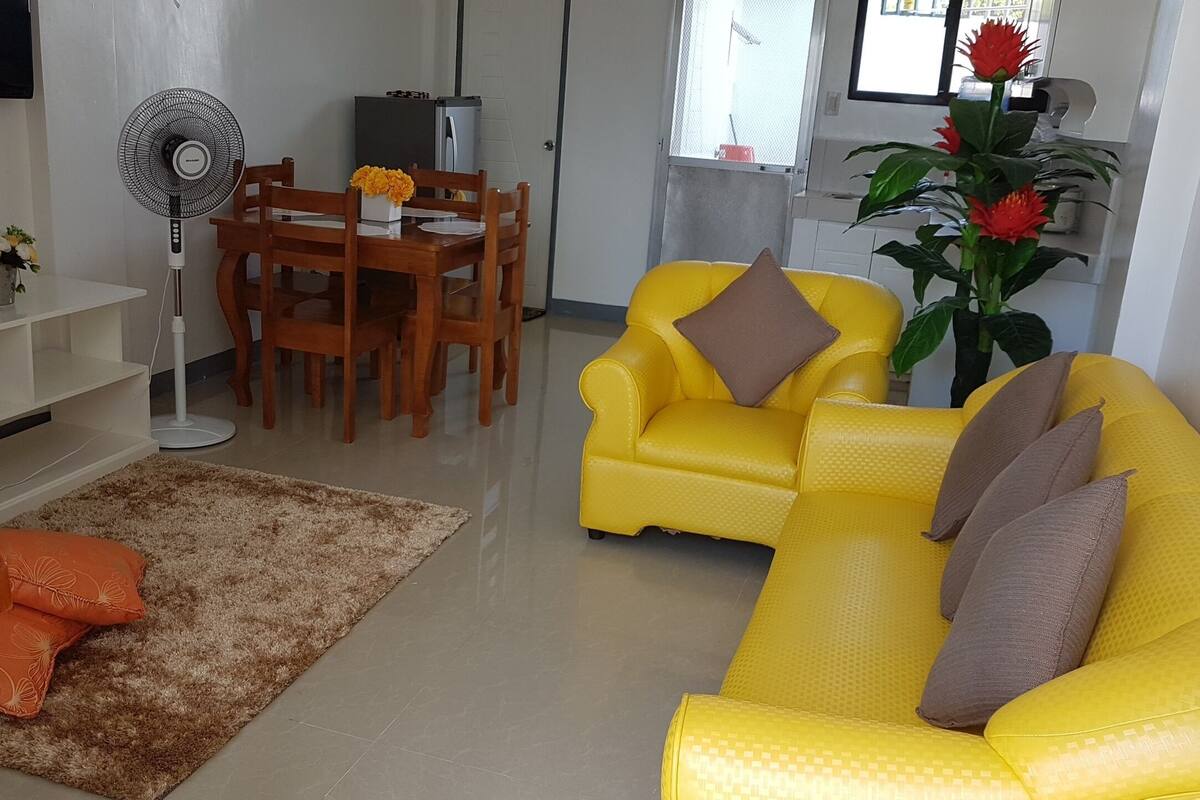A living room with a yellow sofa, a brown armchair, a dining table with chairs, a standing fan, and a potted plant.