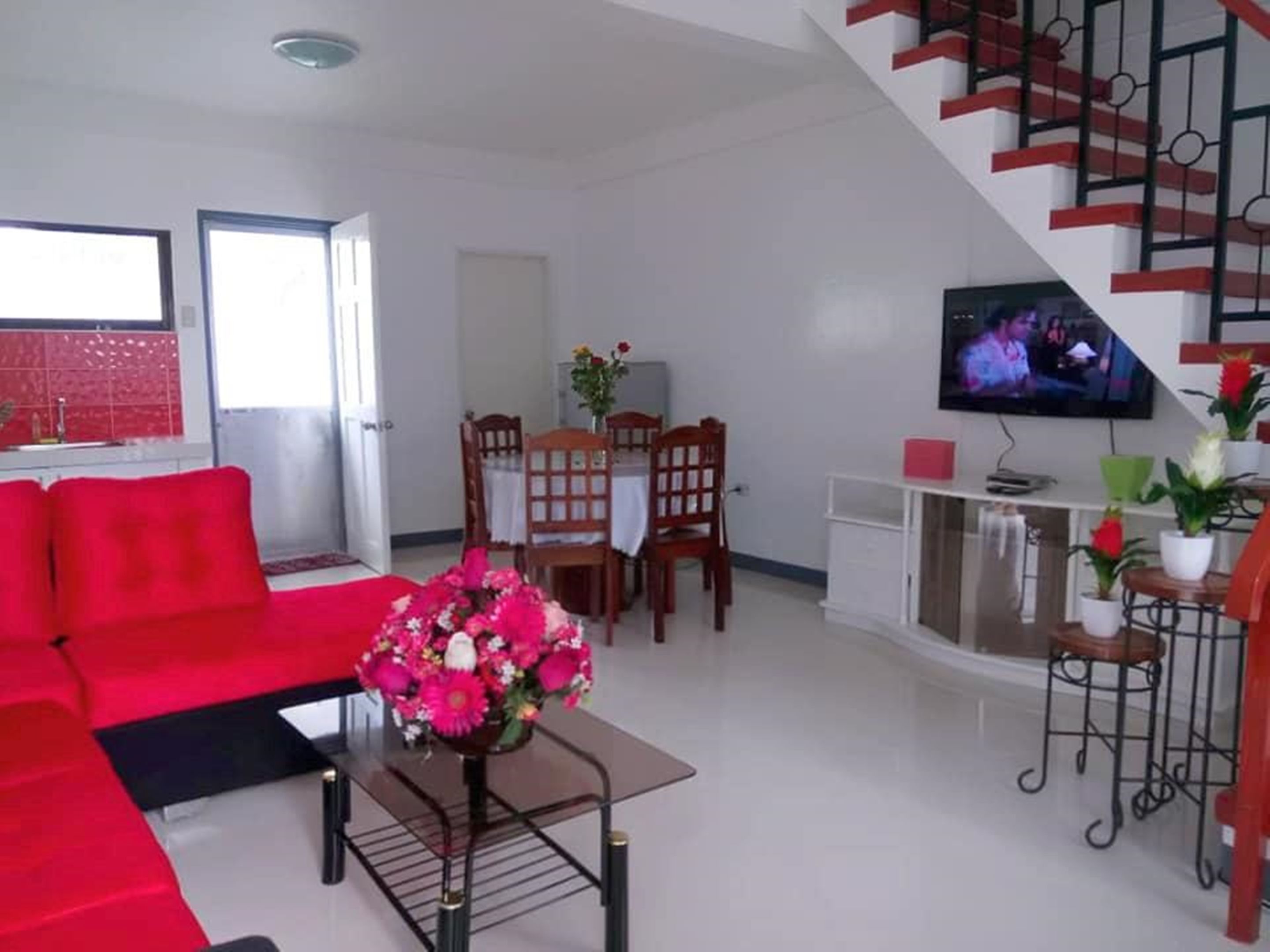 A living room with a red sofa, a coffee table with a floral arrangement, a dining area with wooden chairs, and a TV on the wall.