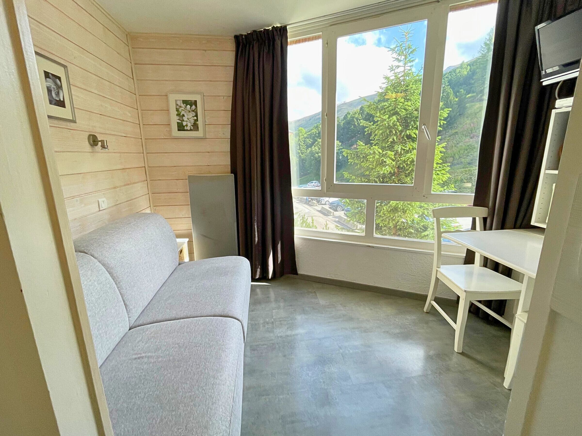 A room with a grey sofa, a small white table, a TV, and a window with curtains.