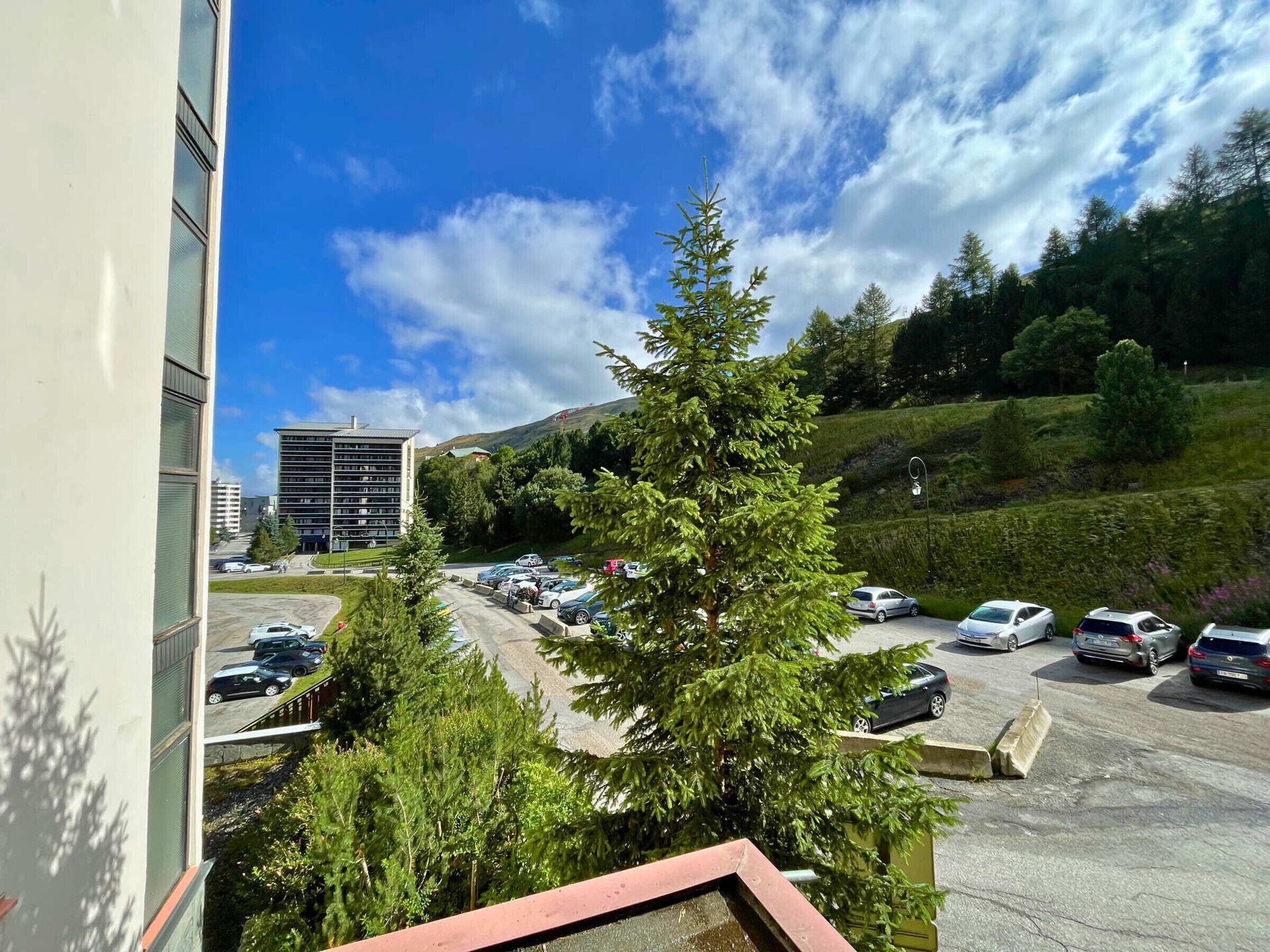 A parking lot with cars, a building with multiple floors, and a hill with trees in the background.