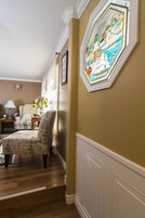 Una habitación con piso de madera, una silla con estampado de la Torre Eiffel, una lámpara y un ventanal de vidrio emplomado montado en la pared.