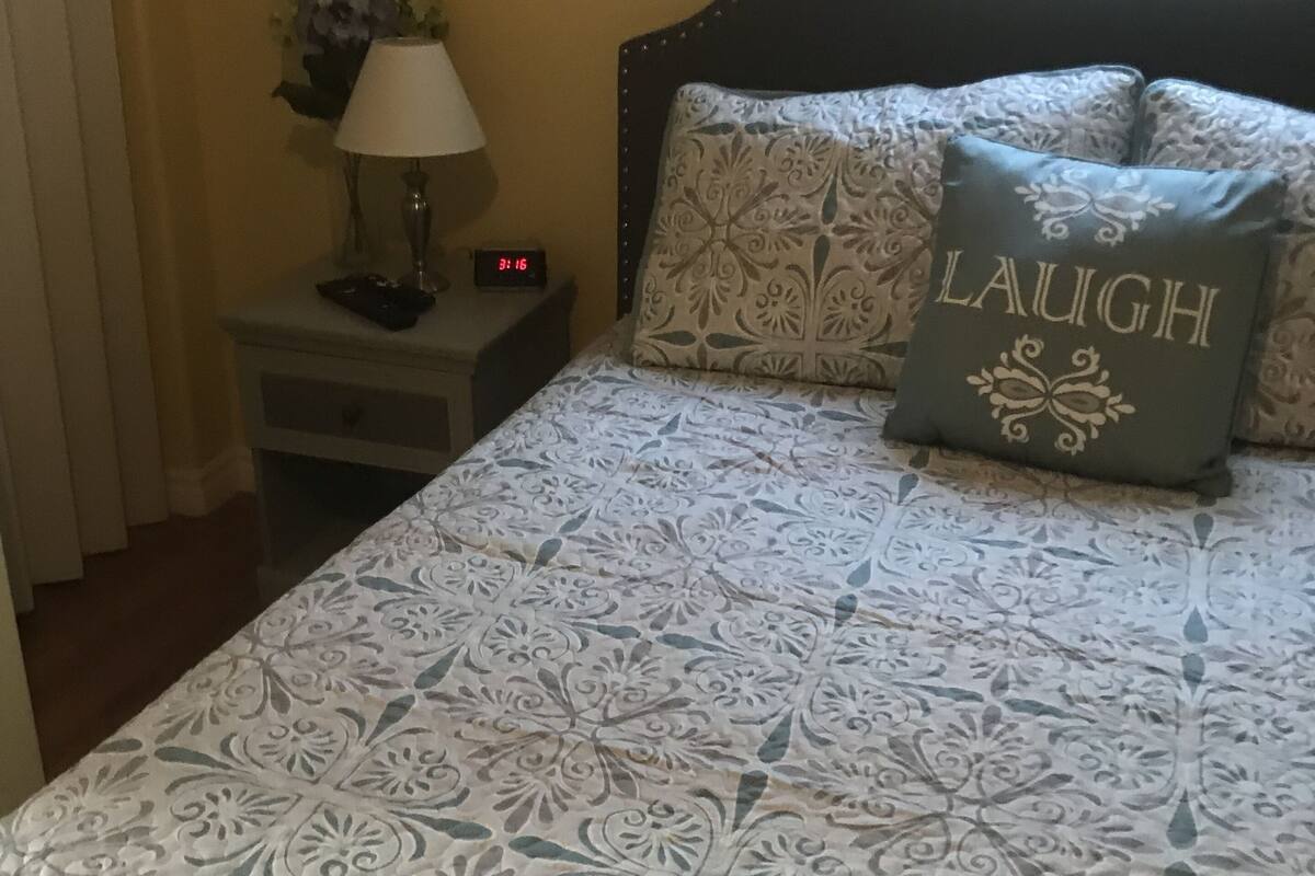 Uma cama bem arrumada com roupa de cama estampada, uma mesa de cabeceira com abajur, um relógio digital e um travesseiro decorativo com a palavra 'LAUGH'.