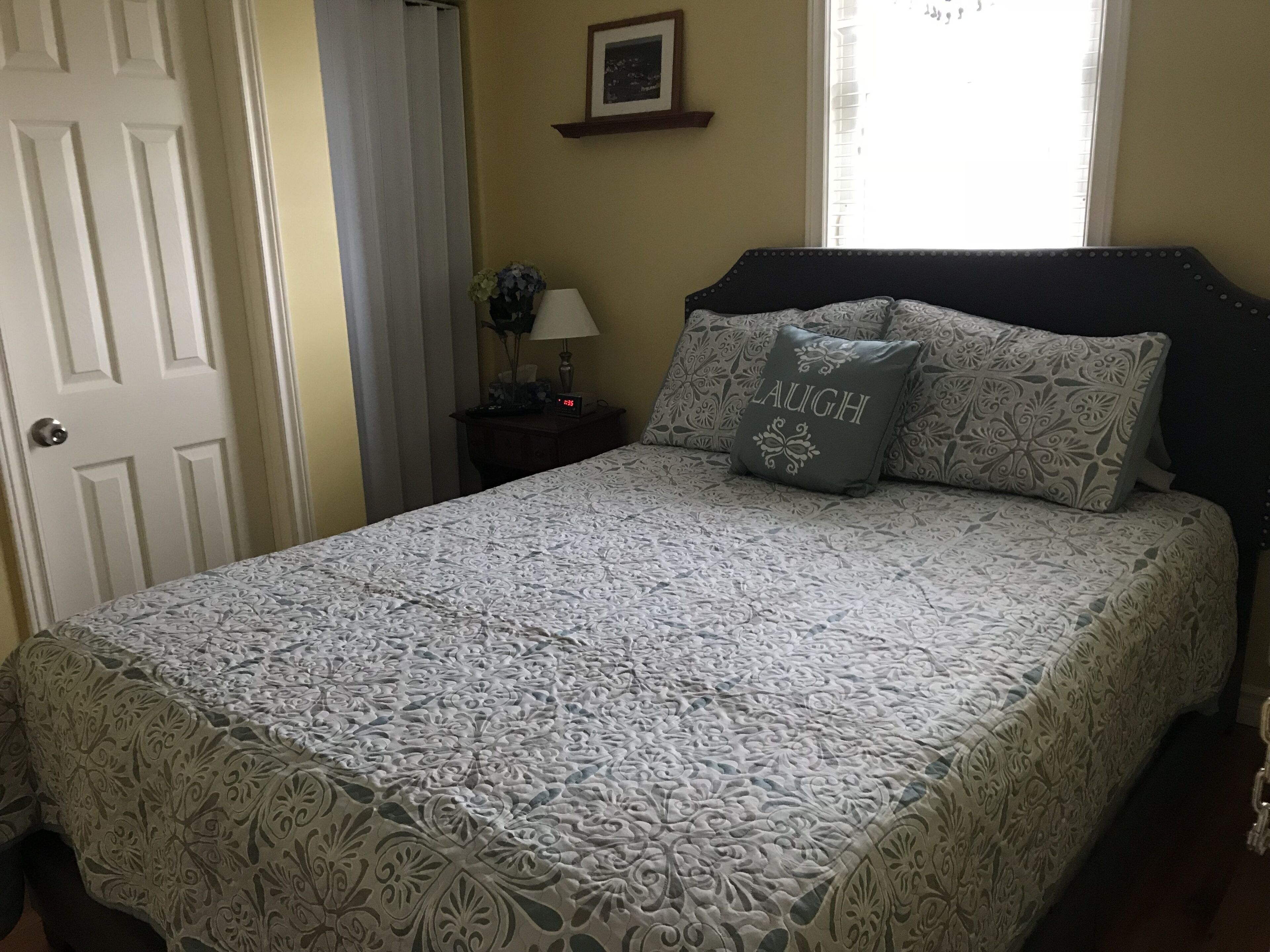 Een net opgemaakt bed met beddengoed met patroon, een nachtkastje met een lamp, en een ingelijst foto aan de muur.