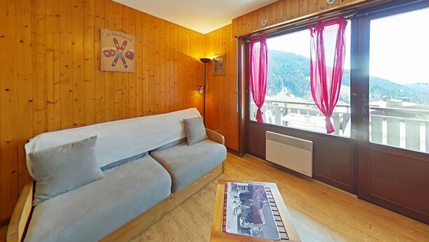 Une pièce chaleureuse avec un mur en bois, un canapé, une table sur laquelle est posée une image et une fenêtre avec des rideaux.