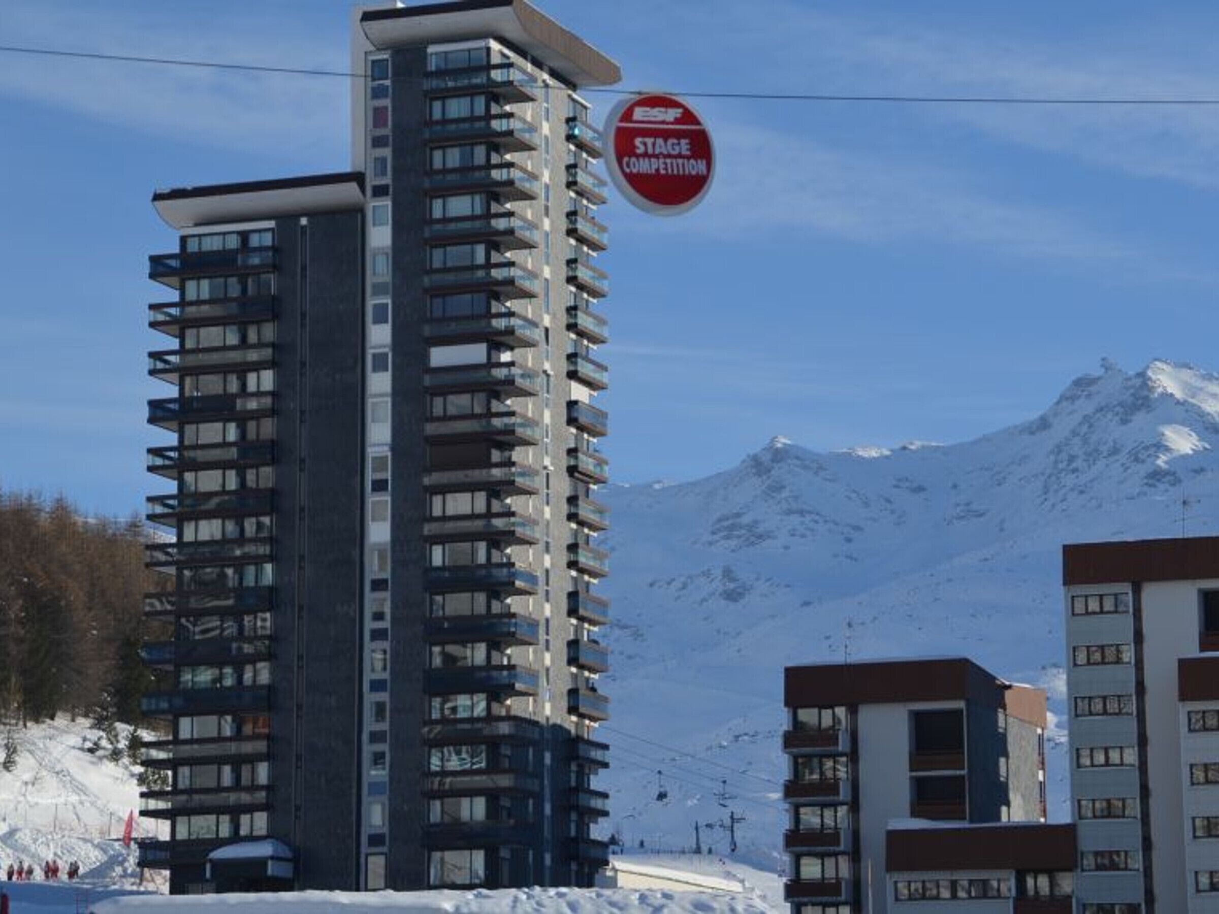 A tall, modern building with multiple balconies, a ski lift in the background, and a snow-covered mountain.