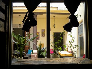 A view through a window with curtains, featuring a wooden table, potted plants, and a glimpse of a building with a tiled roof and a hanging lantern. - Casa I'X - Hostel (Quetzaltenango)