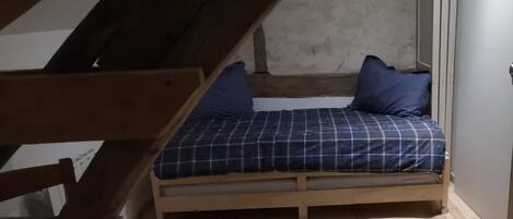 Pieni huone, jossa on yksi sänky, kalteva katto, puulattia ja pelkistetty sänky sinisillä vuodevaatteilla.