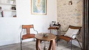 Una habitación con dos sillones de mimbre, una mesa de madera, un cuadro enmarcado, un estante con adornos y una pared de piedra.