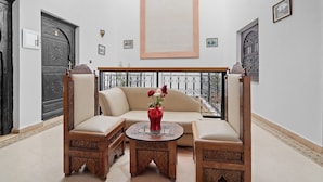 A room with a beige sofa, two wooden chairs, a small round table with a red vase, and a balcony with a wrought iron railing. - Riad Marwa (Marrakech)