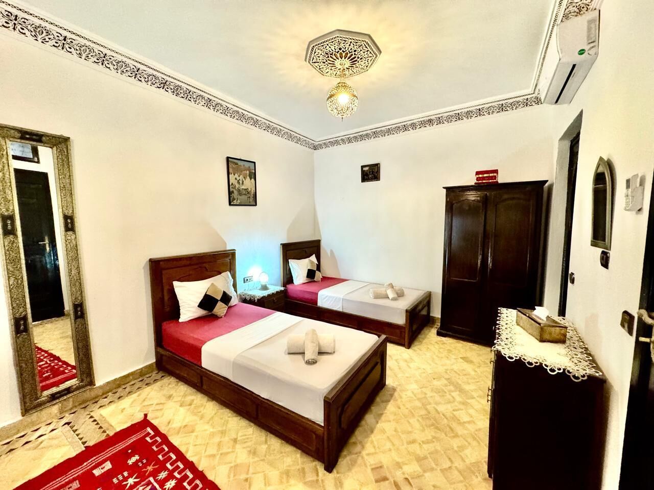 A hotel room with two beds, a wooden wardrobe, a red rug, and a decorative ceiling light fixture.