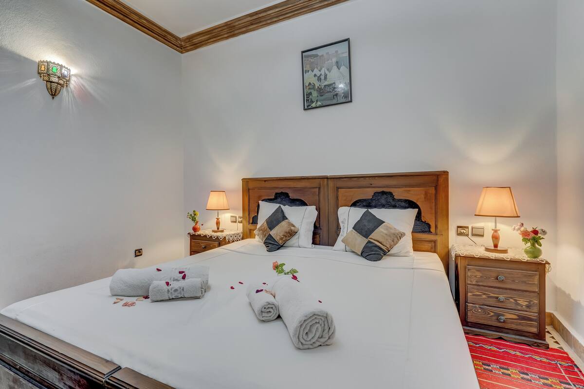 A neatly made bed with rolled towels and rose petals, wooden headboard, two bedside tables with lamps, and a framed picture on the wall.