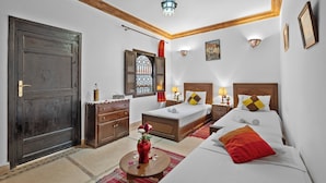 A hotel room with a large wooden door, a bed with white linens, a red throw pillow, a wooden nightstand with a lamp, and a small table with a vase of flowers. - Riad Marwa (Marrakech)