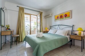A hotel room with a green bedspread, a desk with a chair, a small table with a lamp, and a window with curtains. - Studios Ritsa (Skiathos)