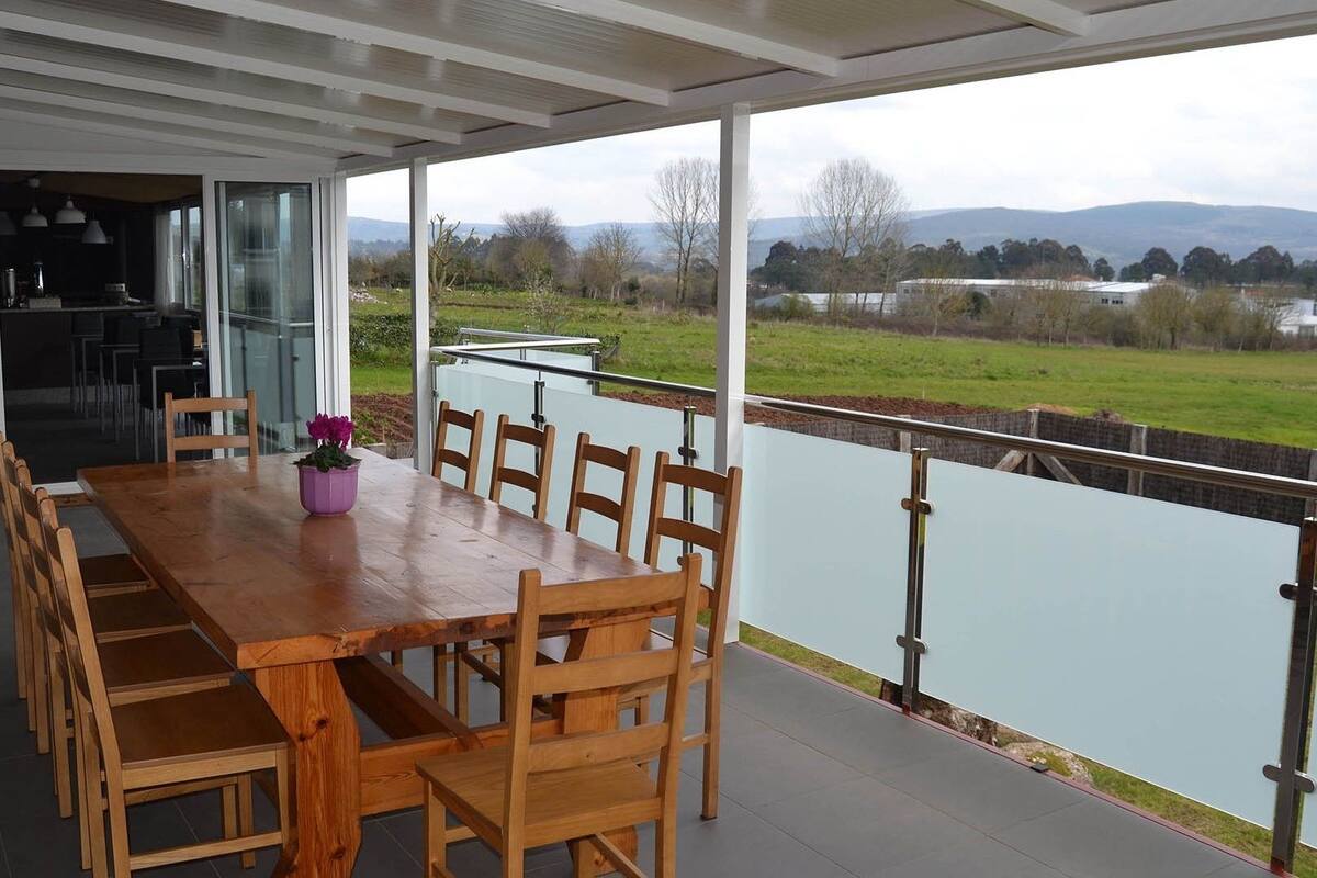 A covered dining area with a wooden table and chairs, a potted plant, and a view of a lake and hills.