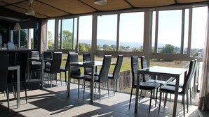 A sunlit dining area with a view of the countryside, featuring a table and chairs under large windows. - Albergue Alfonso II el Casto - Hostel (Melide)