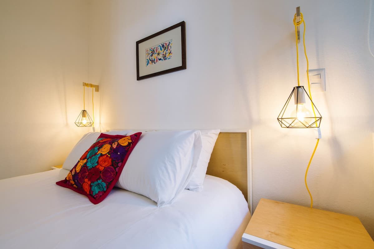 A neatly made bed with white linens and a colorful floral pillow, a framed abstract artwork on the wall, and two wall-mounted lamps with yellow cords.