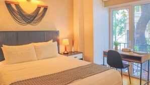 A hotel room with a bed, bedside table, lamp, desk, chair, and a window with a view of greenery. - MyPlace Zócalo (Mexico City)