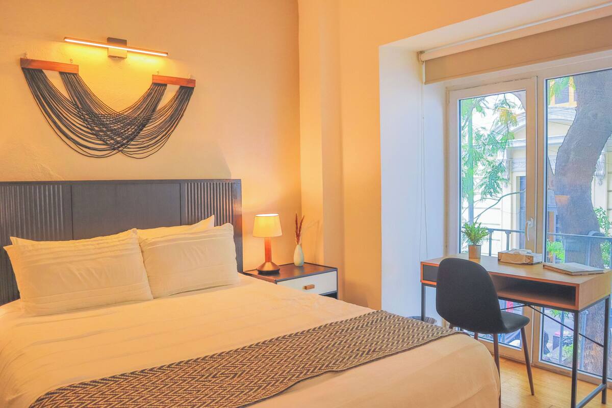 A hotel room with a bed, bedside table, lamp, desk, chair, and a window with a view of greenery.
