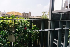 A balcony with a metal railing, lush green plants, and a view of a building with a dome and a tower in the background. - MyPlace Zócalo (Mexico City)