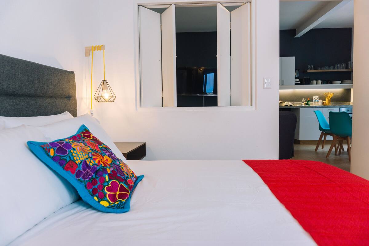 A hotel room with a bed, a colorful pillow, a nightstand, a wall-mounted lamp, a window, and a view into the kitchen area.