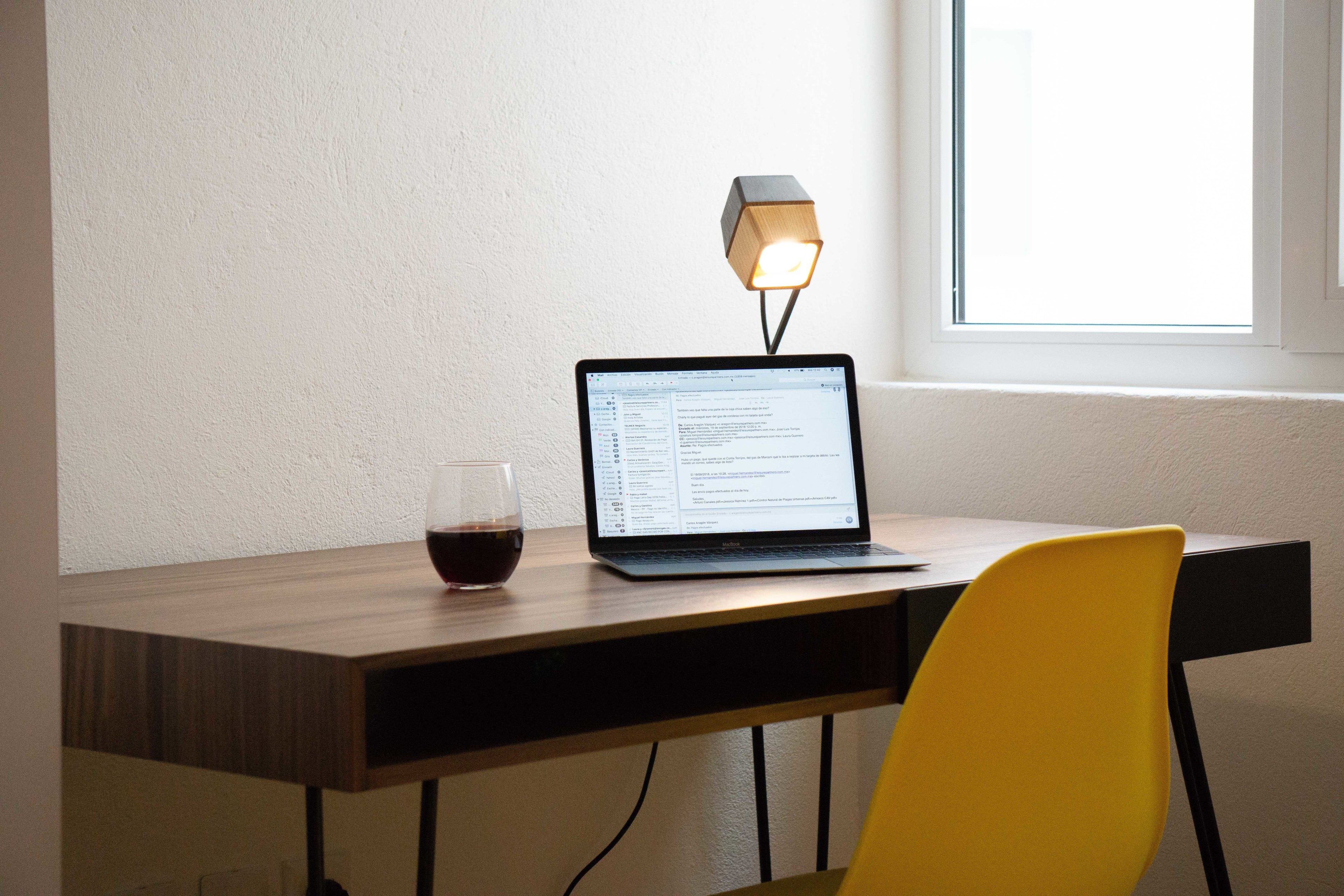 A laptop on a wooden desk with a glass of red wine, a yellow chair, and a wall-mounted lamp.