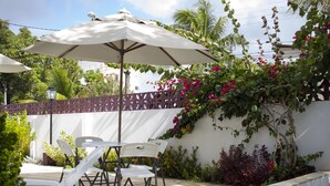 An outdoor patio with white chairs and a table under a large umbrella, surrounded by lush greenery and flowering plants. - Pousada Espaço Viana (Ipojuca)