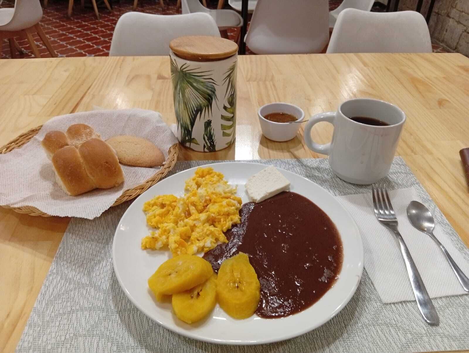 A breakfast setting with a plate of scrambled eggs, beans, and plantains, a cup of coffee, and a basket of bread rolls.