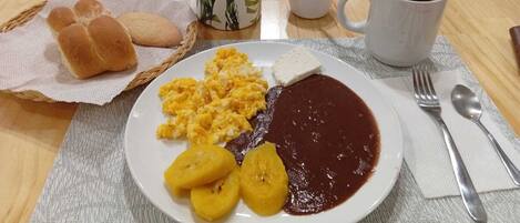 A breakfast setting with a plate of scrambled eggs, beans, and plantains, a cup of coffee, and a basket of bread rolls.