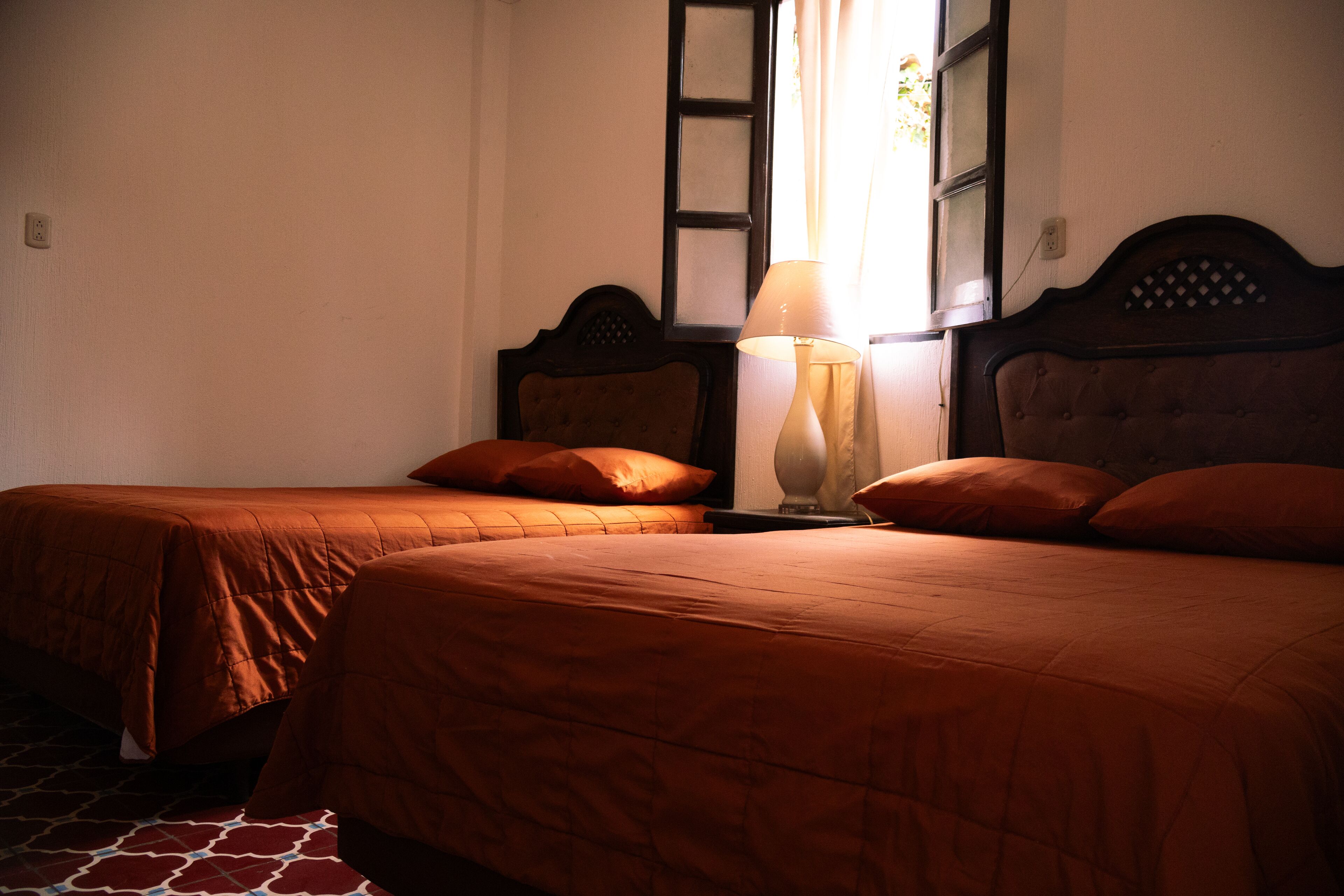 A room with two beds, each with a wooden headboard and matching pillows. There is a nightstand with a lamp between the beds.