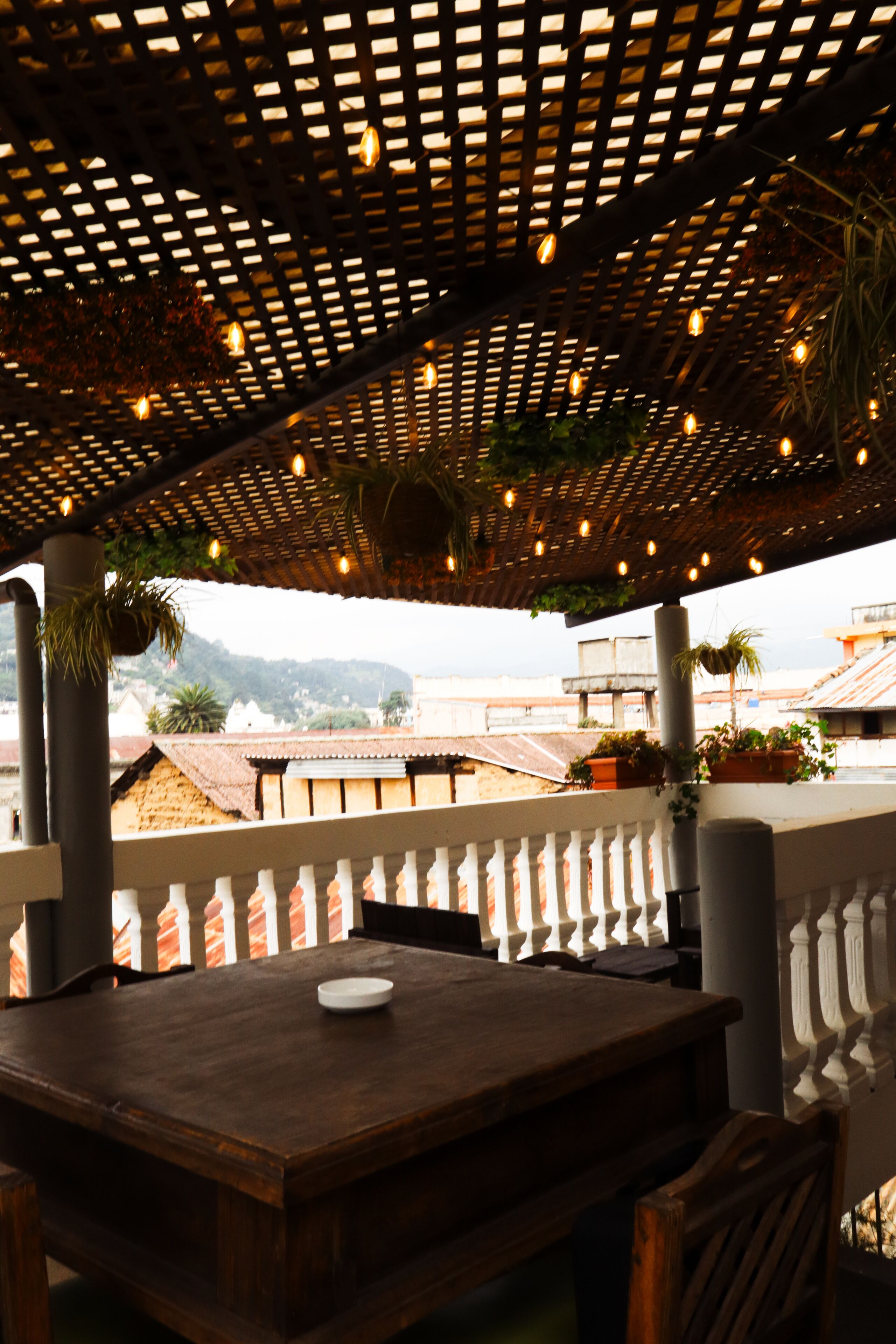 An outdoor patio with a wooden table and chairs, a white balustrade, hanging plants, and a view of buildings and hills in the background.