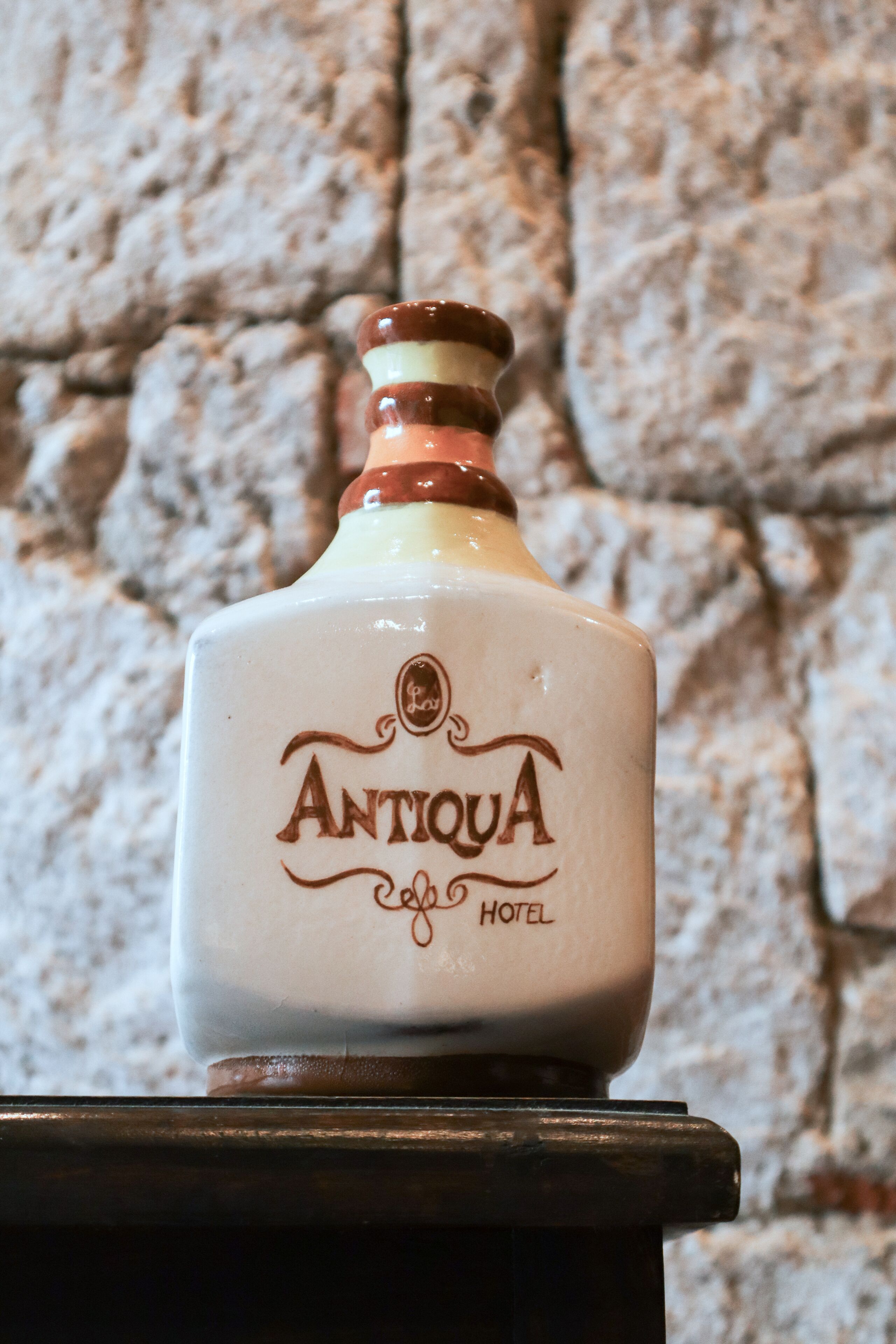 Una ampolla amb l'etiqueta “Antigua Hotel” sobre una superfície de fusta, amb un mur de pedra de fons.