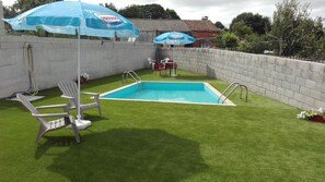 A swimming pool with artificial grass, two white plastic chairs, and two blue umbrellas.