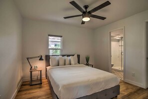 A bedroom with a bed, a ceiling fan, a nightstand with a lamp, a window, and a door leading to a bathroom.