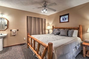 A bedroom with a wooden bed, a ceiling fan, a window with curtains, a bedside lamp, and a bathroom with a sink and mirror. - Gatlinburg Home w/ Hot Tub ~ 4 Mi to Town! (Gatlinburg)