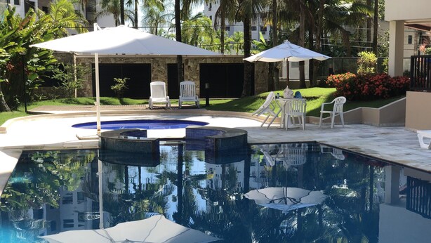 A swimming pool with a hot tub, surrounded by white chairs and umbrellas, with palm trees and a stone building in the background. - Riviera apartment, 50m from the beach 3 bedrooms 2bh, living room 2 rooms, balcony, air conditioning, wi-fi  (Bertioga)