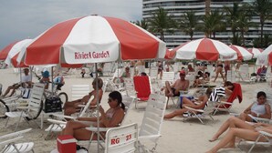 A beach with white and red striped umbrellas, white lounge chairs, and a large building in the background. - Riviera apartment, 50m from the beach 3 bedrooms 2bh, living room 2 rooms, balcony, air conditioning, wi-fi  (Bertioga)