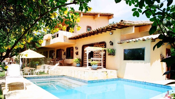A pool area with lounge chairs, a pergola, and a building with a tiled roof.