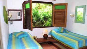 A room with two beds, an air conditioning unit, and a window with a view of greenery.