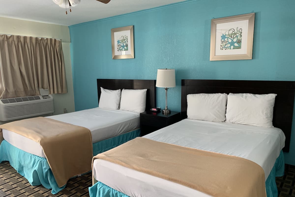 Une chambre d’hôtel avec deux lits, une table de chevet avec une lampe, un climatiseur mural et deux œuvres d’art encadrées accrochées au mur.