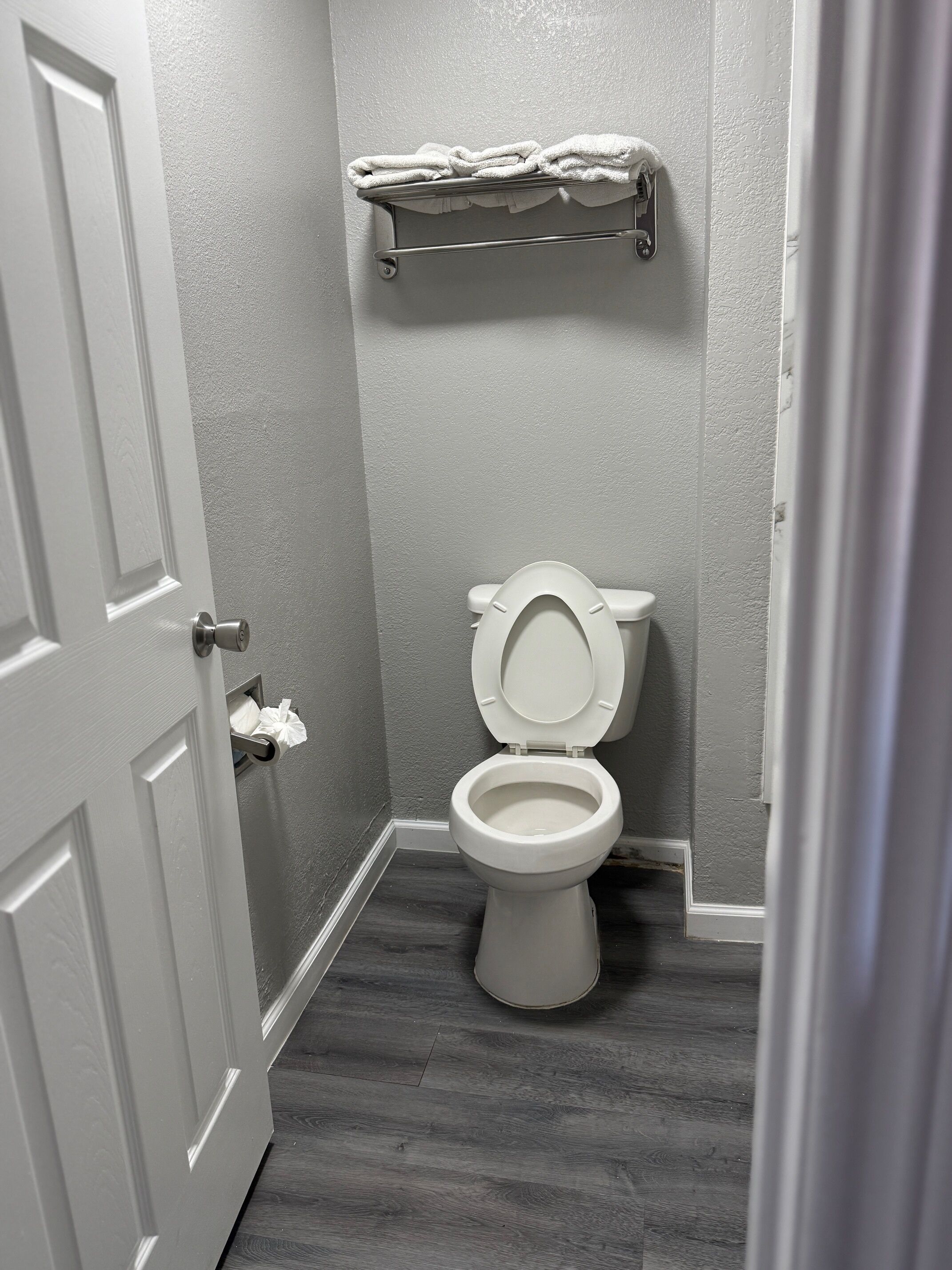 A bathroom with a toilet, a towel rack with folded towels, and a door with a towel dispenser.