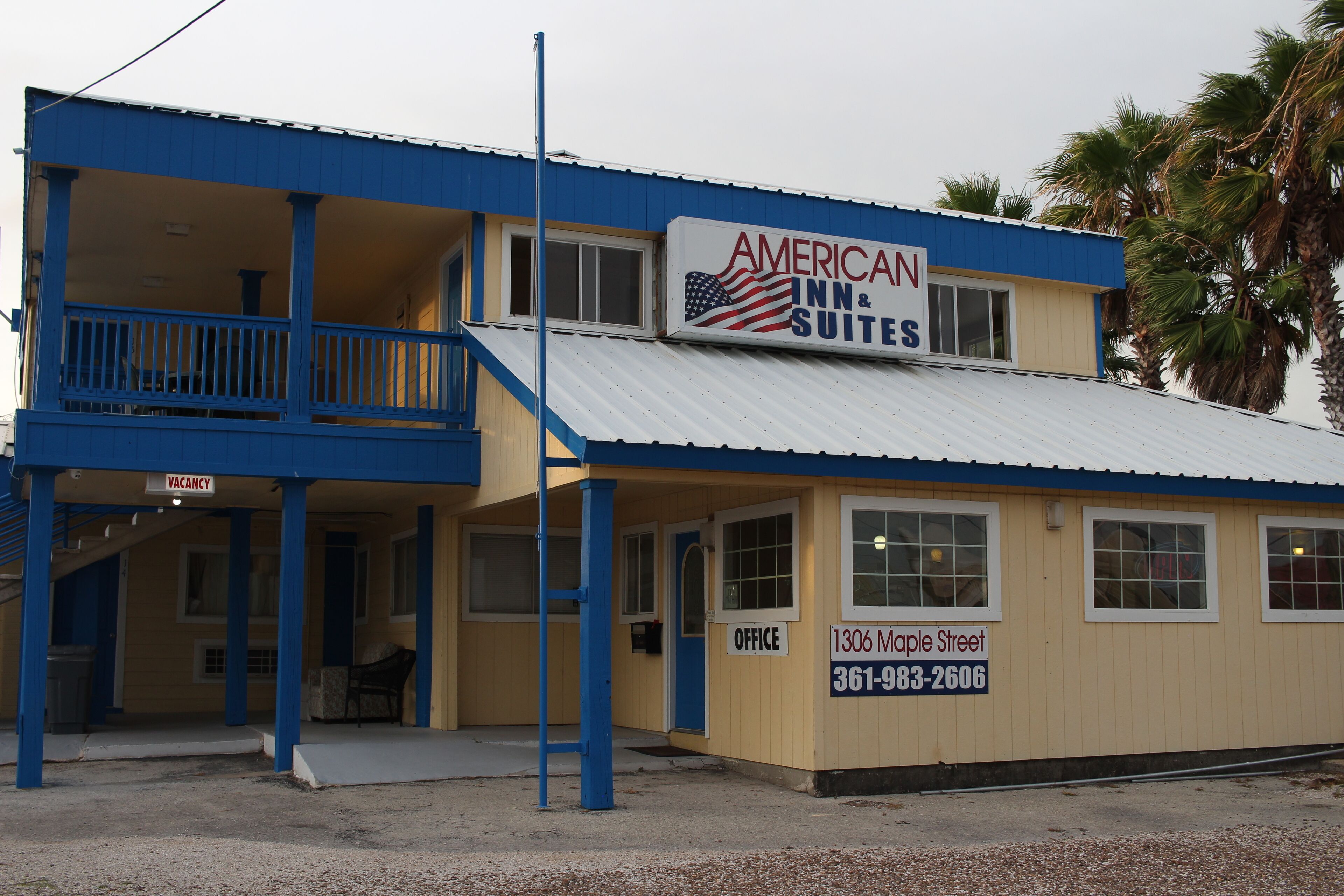 A two-story building with a blue and beige color scheme, featuring a sign for American Inn & Suites, a visible office, and a staircase leading to a balcony.