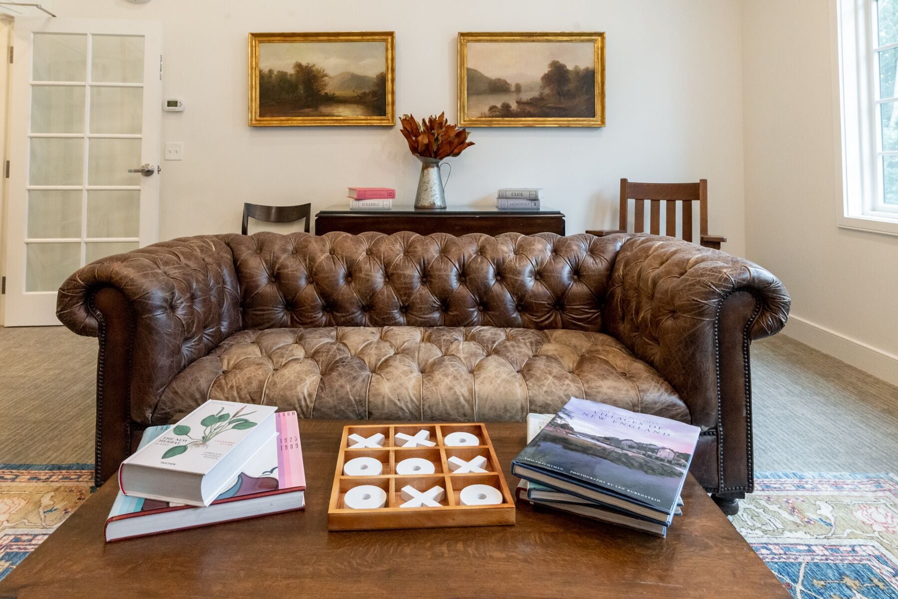Un sofá de cuero marrón en una habitación con dos cuadros enmarcados en la pared, una mesita de madera con libros y un cofre de madera, y una alfombra estampada.