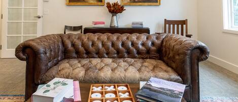 A brown leather sofa in a room with two framed paintings on the wall, a wooden side table with books and a wooden box, and a patterned rug.