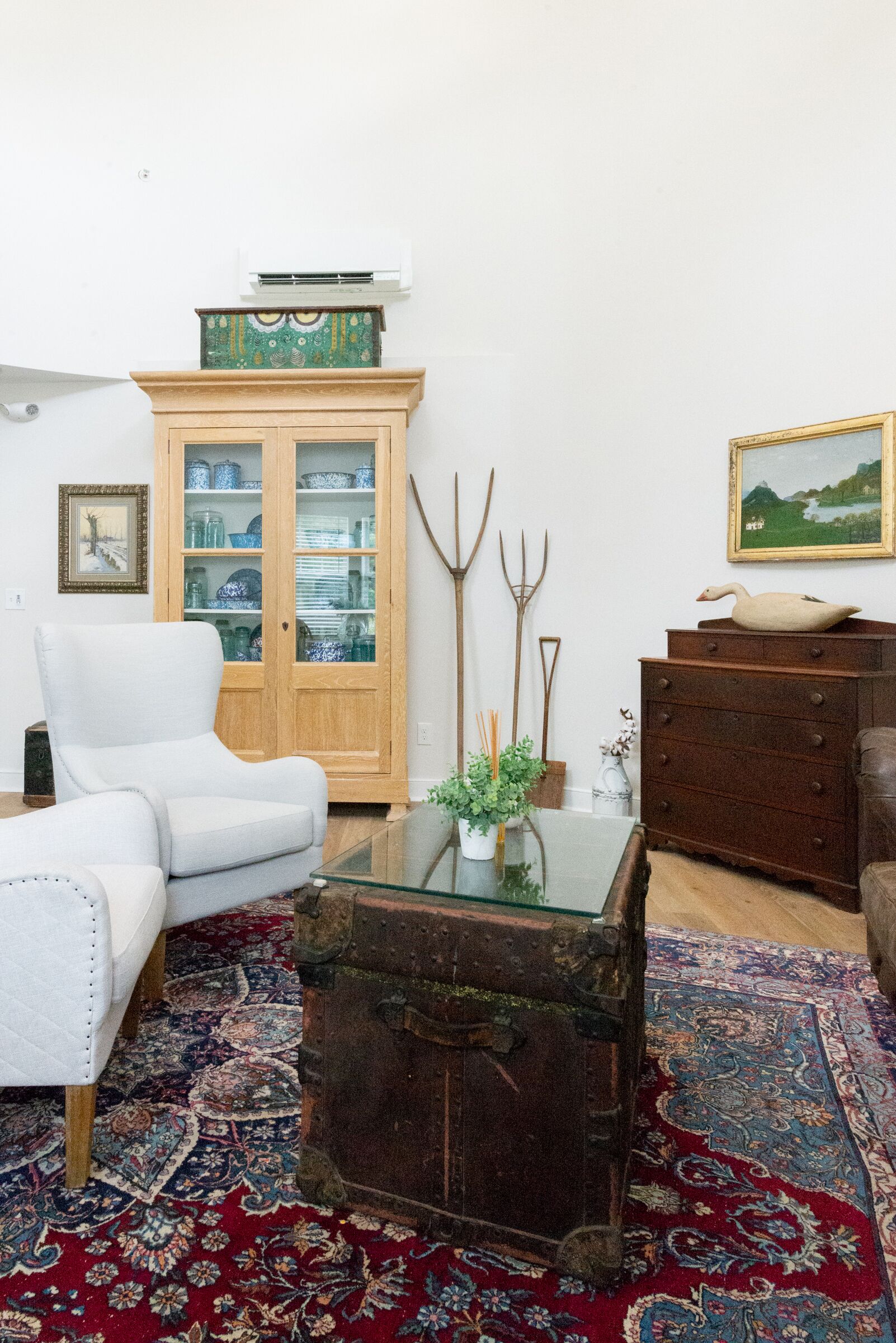 A living room with a white armchair, a wooden cabinet with glass doors, a rustic chest of drawers, and a patterned rug.