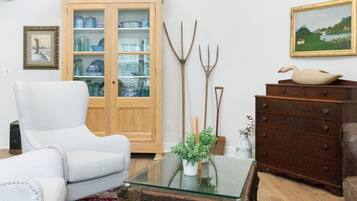 A living room with a white armchair, a wooden cabinet with glass doors, a rustic chest of drawers, and a patterned rug.