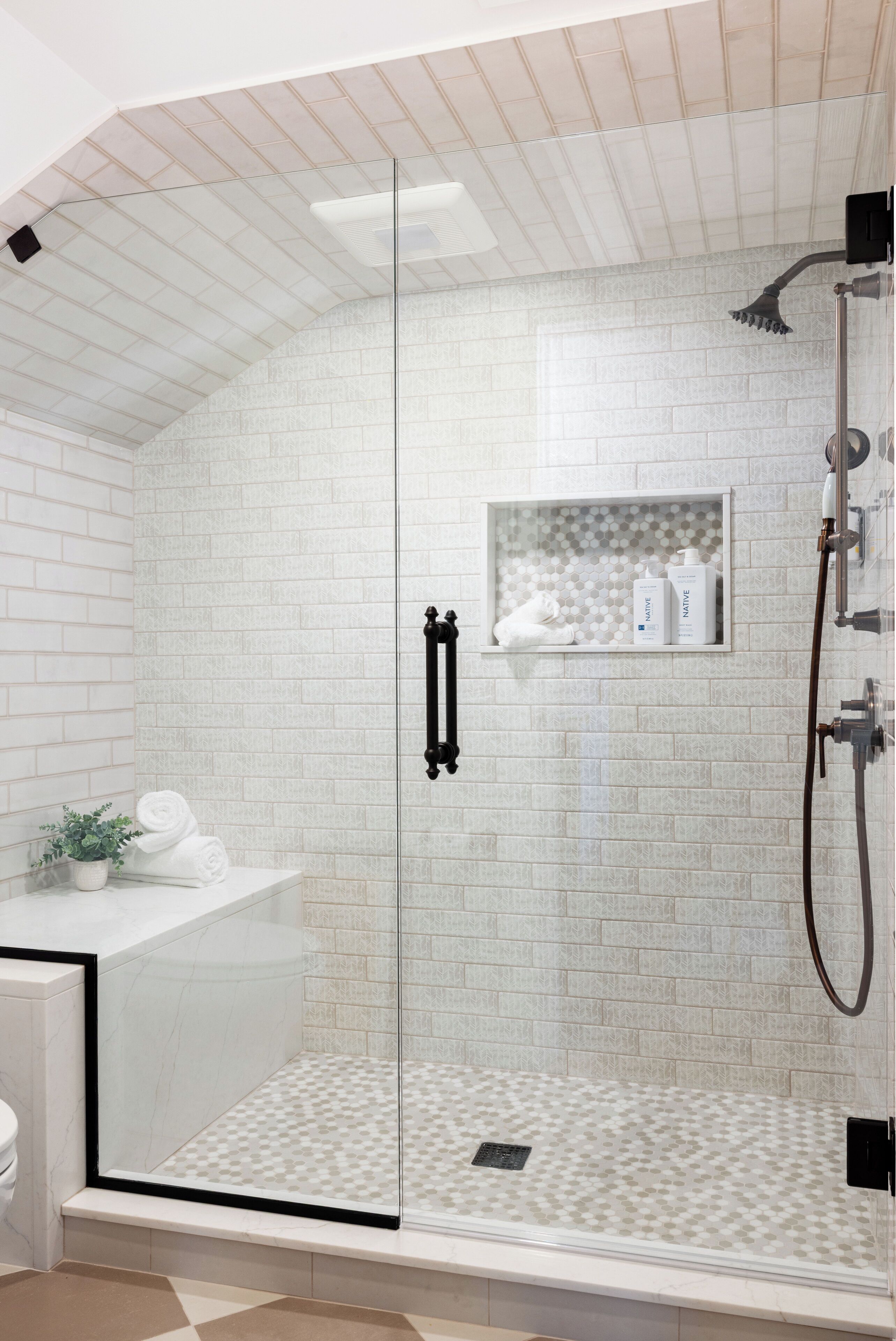 A modern bathroom with a glass-enclosed shower, white tiled walls, and a small shelf with toiletries.