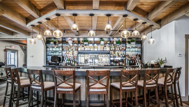 Zona de bar con vigas de madera en el techo, lámparas colgantes y una barra repleta de diferentes botellas. Hay seis taburetes de madera con asientos acolchados.