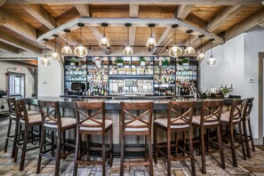 A bar area with wooden ceiling beams, hanging pendant lights, and a bar stocked with various bottles. There are six wooden bar stools with cushioned seats. - Briar Barn Inn (Rowley)