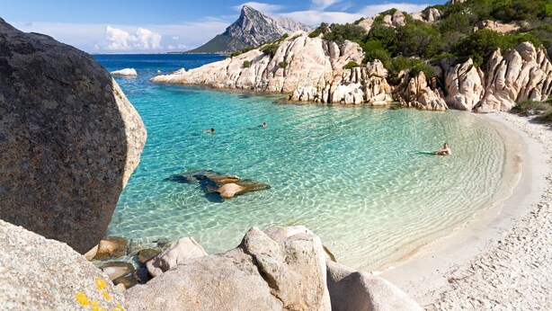 A coastal beach with clear turquoise waters, rocky formations, and a mountainous backdrop.