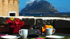 A table with a coffee pot, two cups, and fruit, overlooking a coastal view with a mountain and a building.